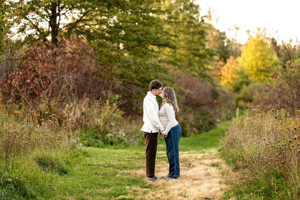 Couple holding hands and kissing on a grassy path surrounded by colorful autumn foliage during an outdoor engagement session.
