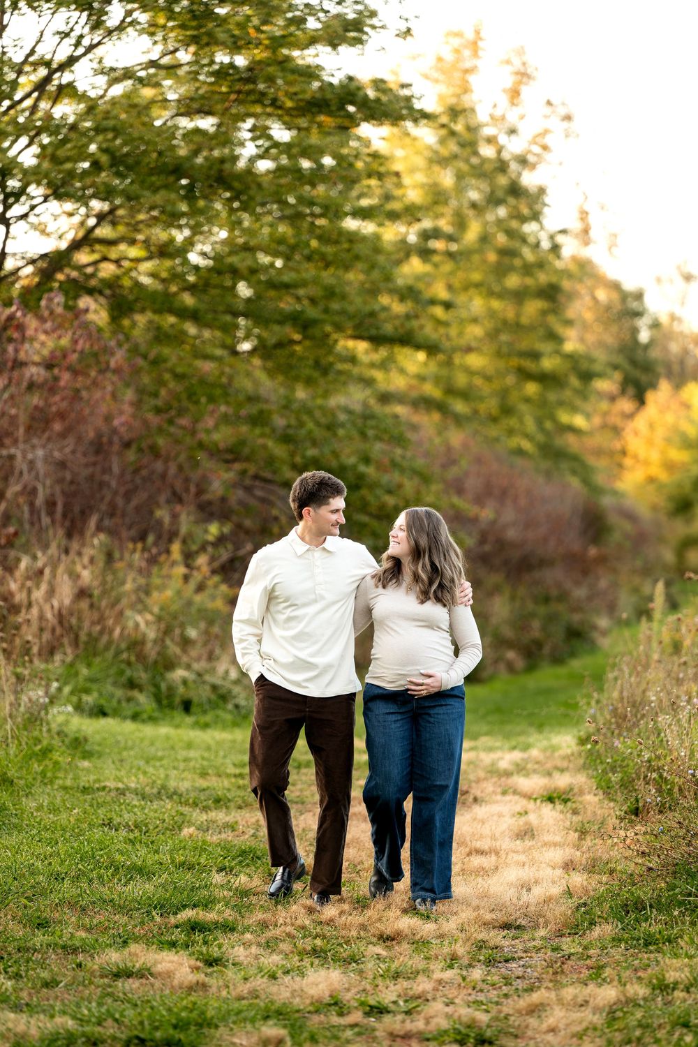 A couple walks along a grassy path surrounded by autumn trees, the woman cradling her pregnant belly.