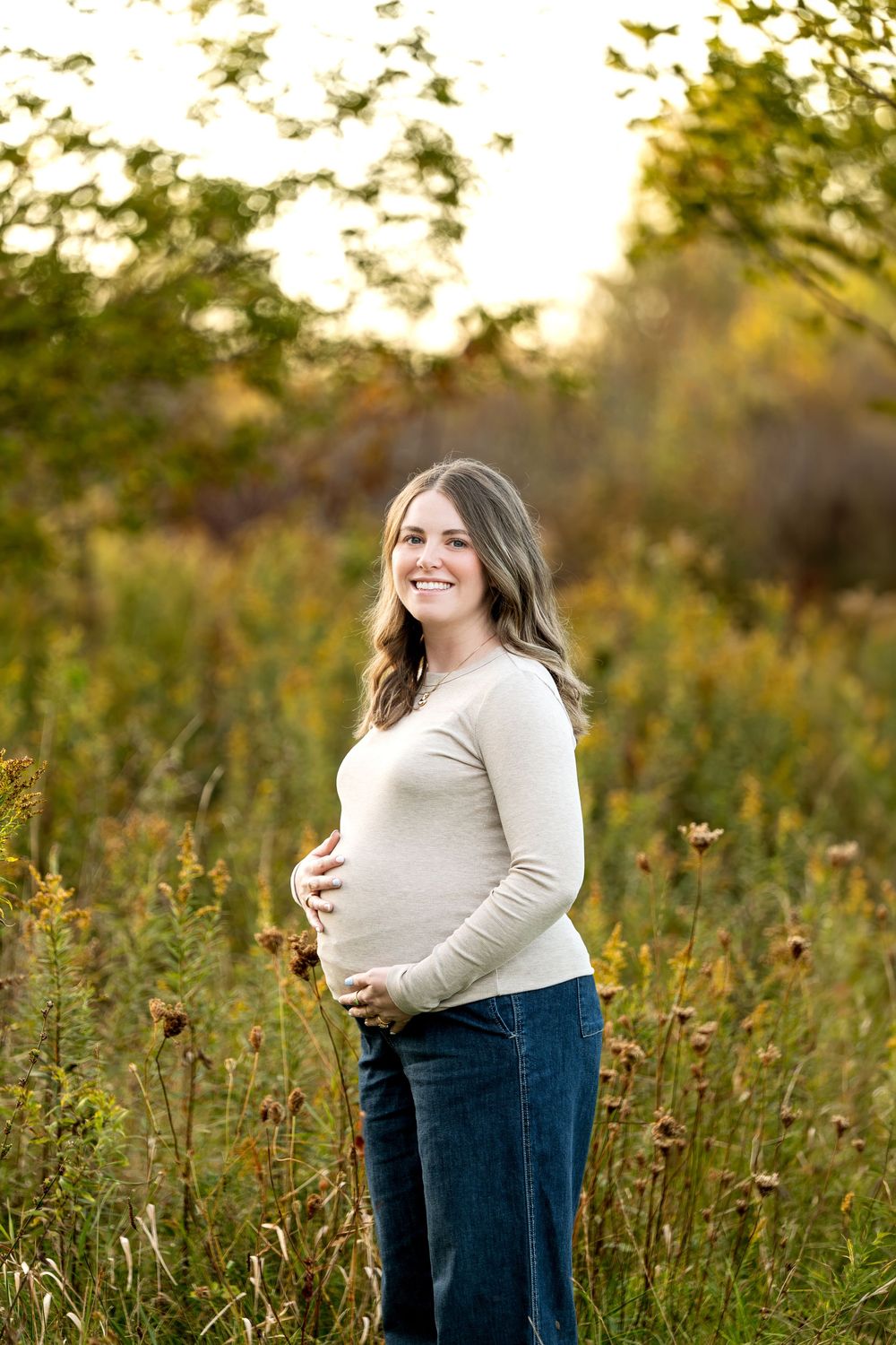Smiling pregnant woman in beige top and jeans cradling baby bump in golden hour field.