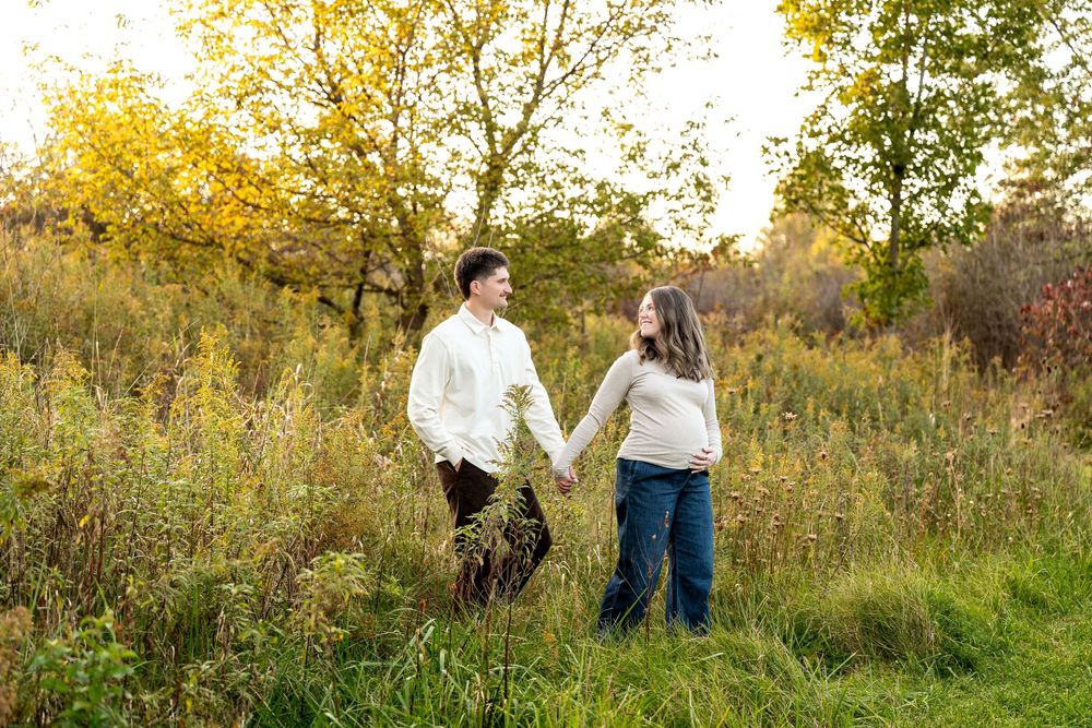 Pregnant couple holding hands in a golden autumn field, smiling at each other during an outdoor maternity photo session.