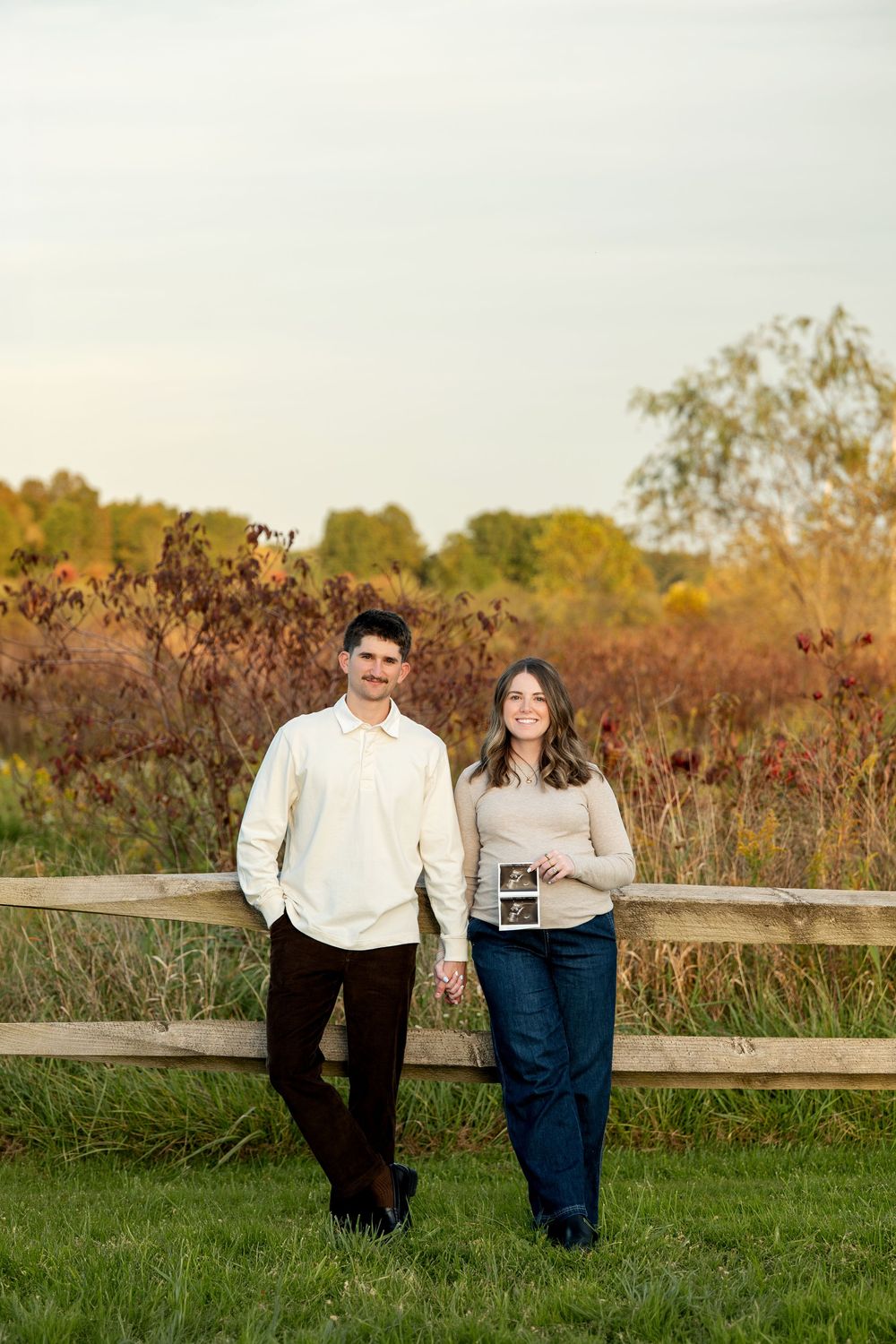 Couple holding ultrasound photo by wooden fence in autumn field, announcing pregnancy outdoors.