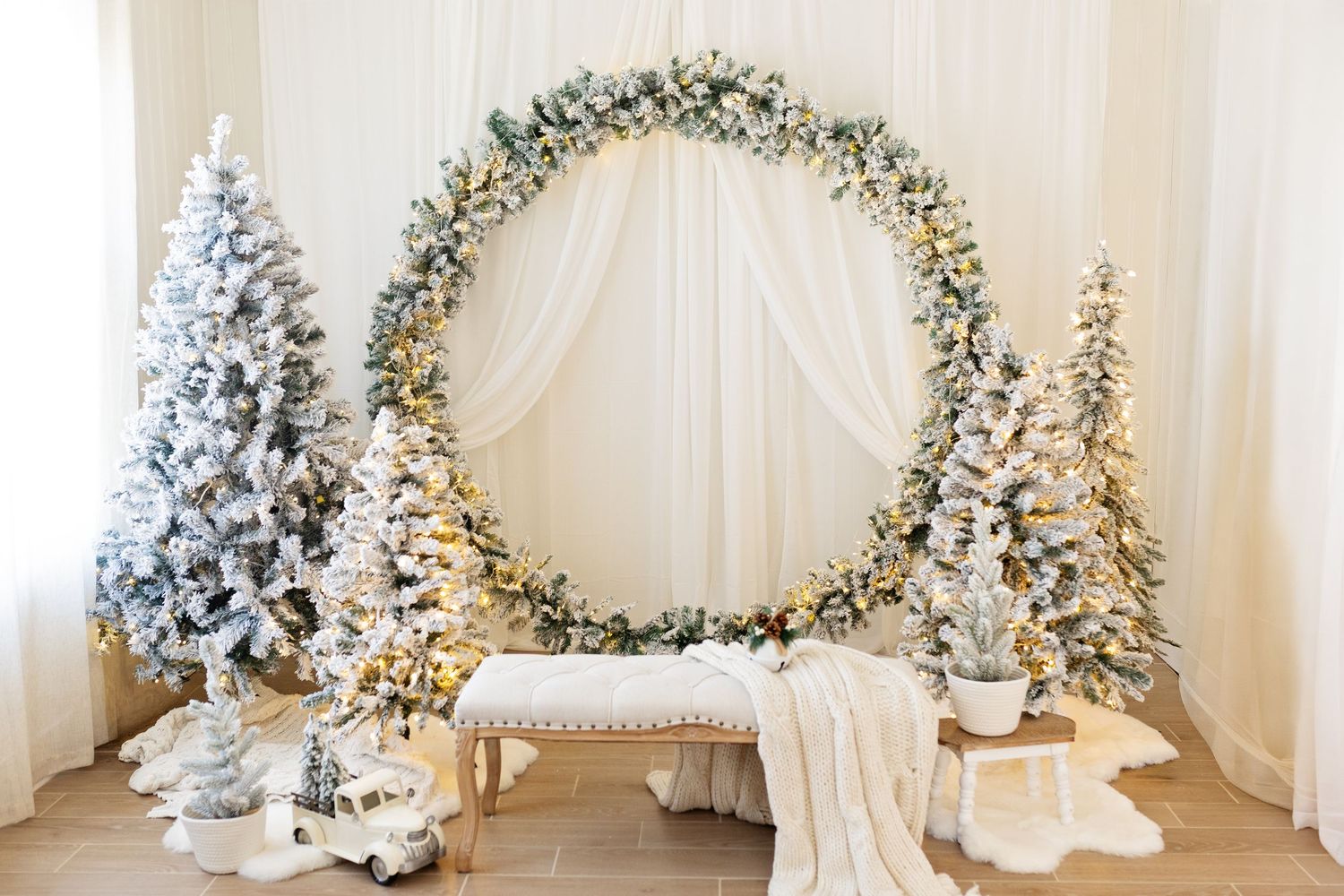 Elegant Christmas photo backdrop with snowy flocked trees, circular wreath, fairy lights, and white draping in a festive studio setting.