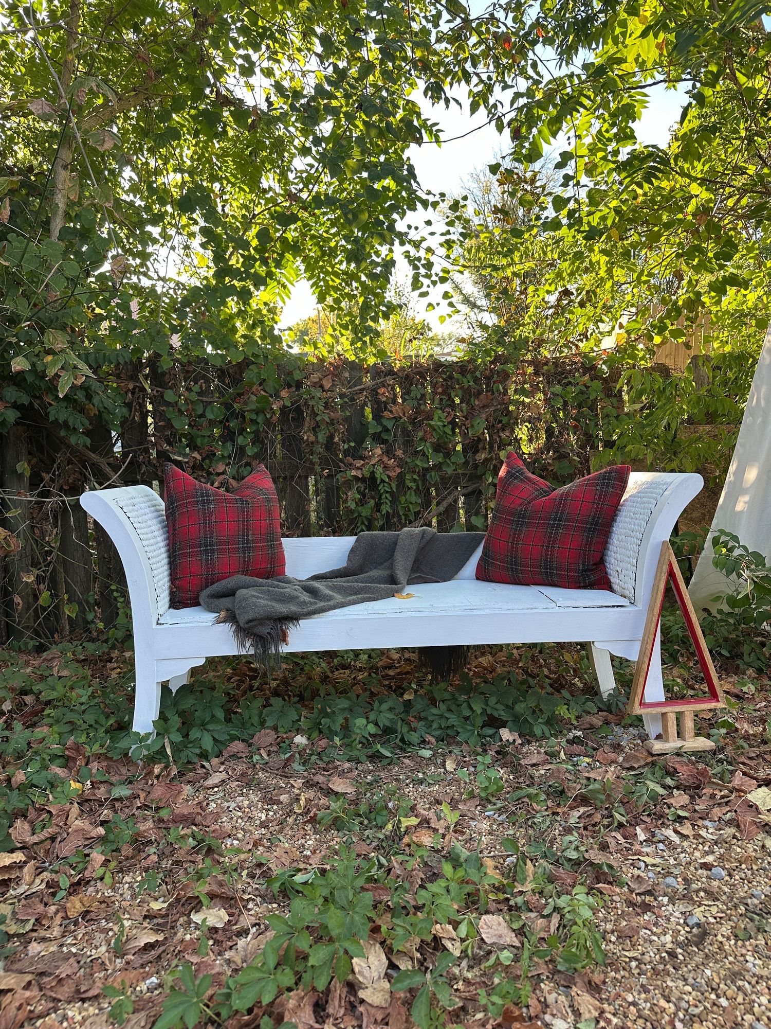White wicker outdoor daybed with red plaid throw pillows and gray blanket, set in a lush garden.