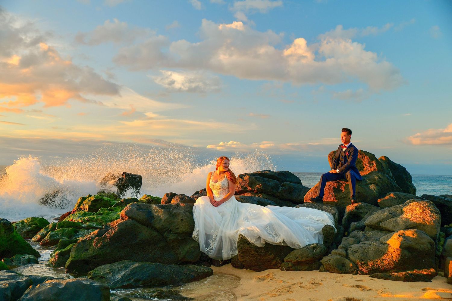 Bride and groom sit on mossy rocks at sunset with crashing waves behind them in a romantic beach wedding photo.