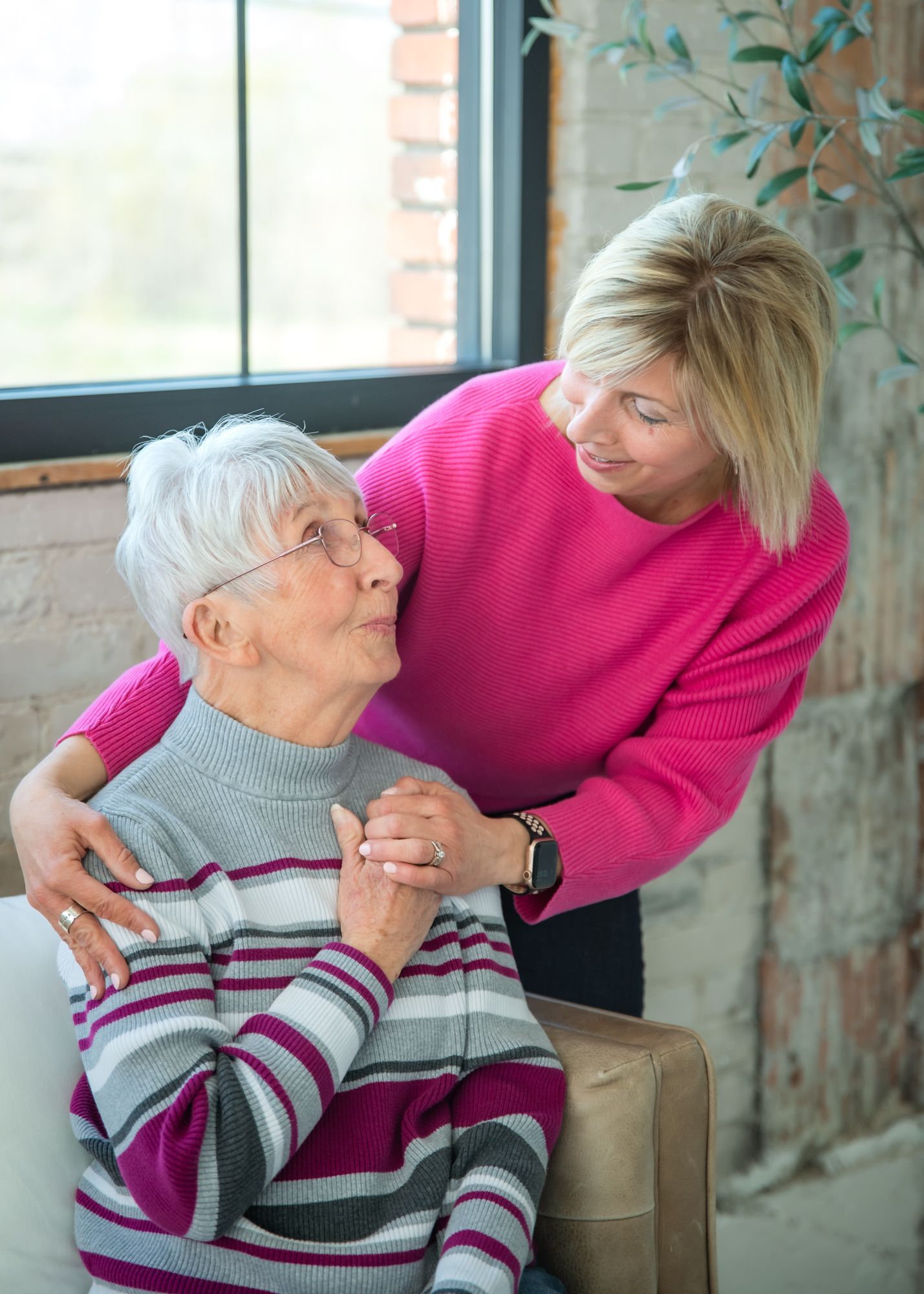Blonde woman in pink sweater tenderly holds hands with elderly white-haired woman in striped top near window.