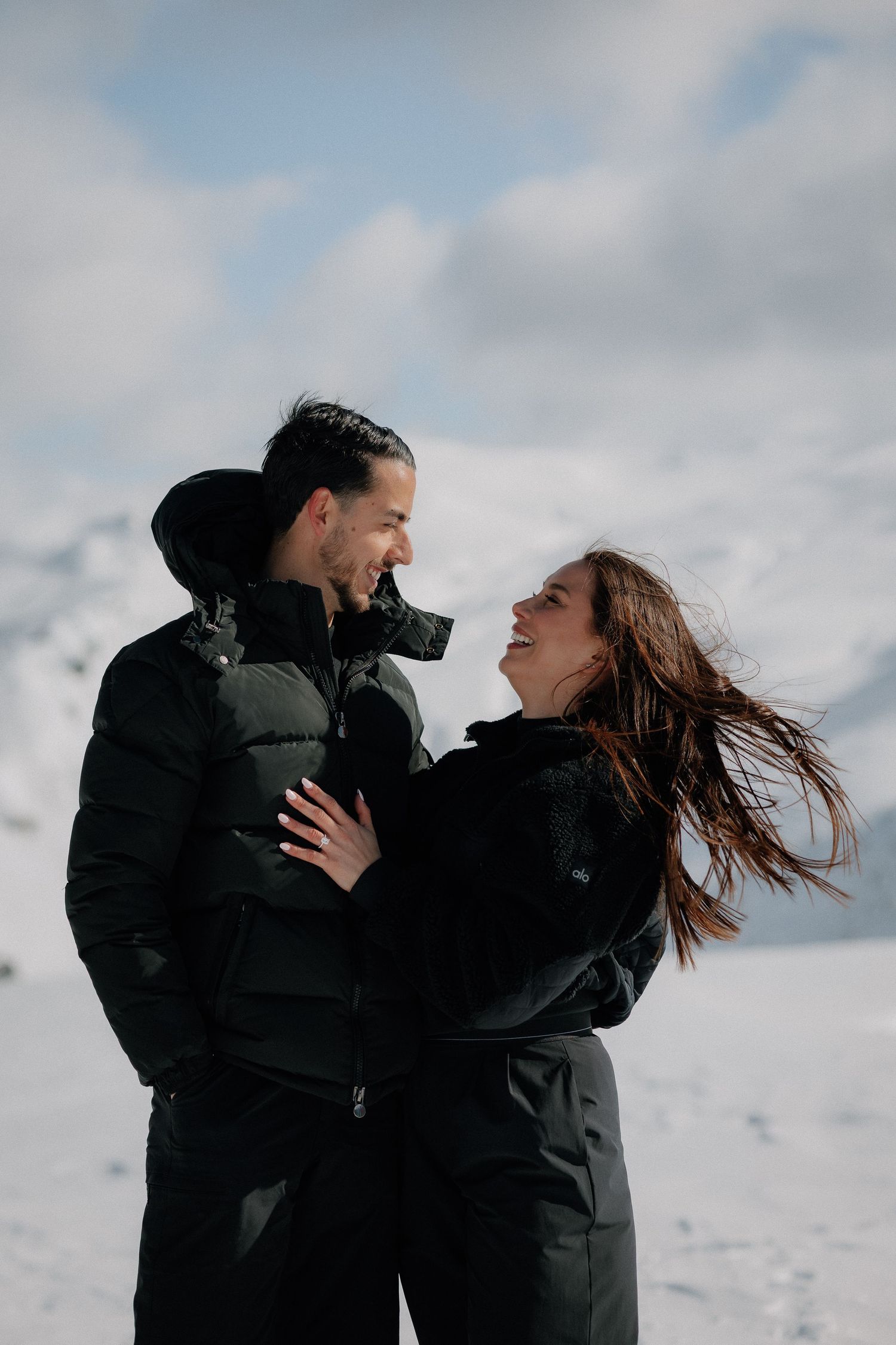 Couple in black winter jackets laughing together on a snowy mountain backdrop with cloudy sky.