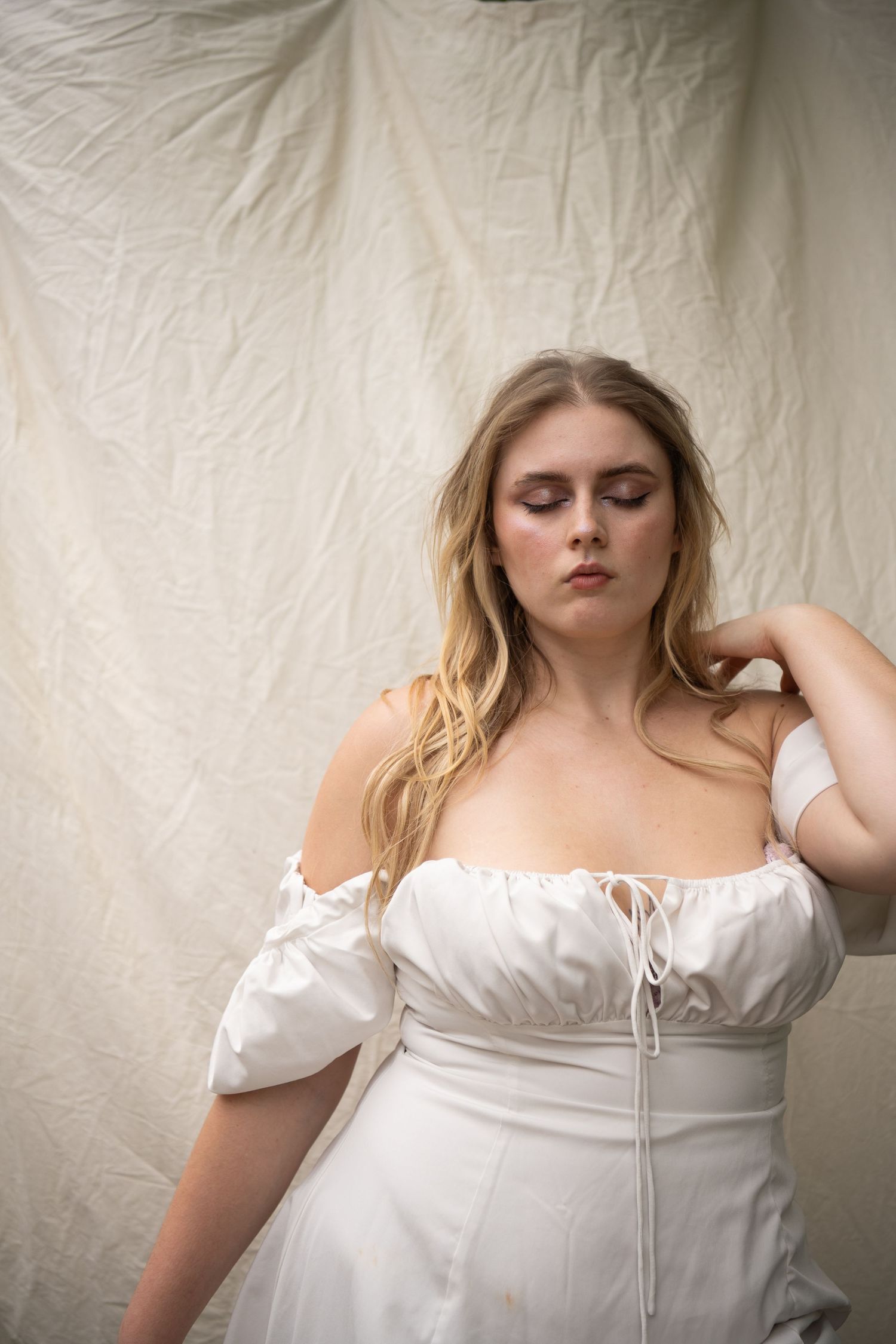 A blonde woman in a white off-shoulder corset dress poses gracefully against a white fabric backdrop.