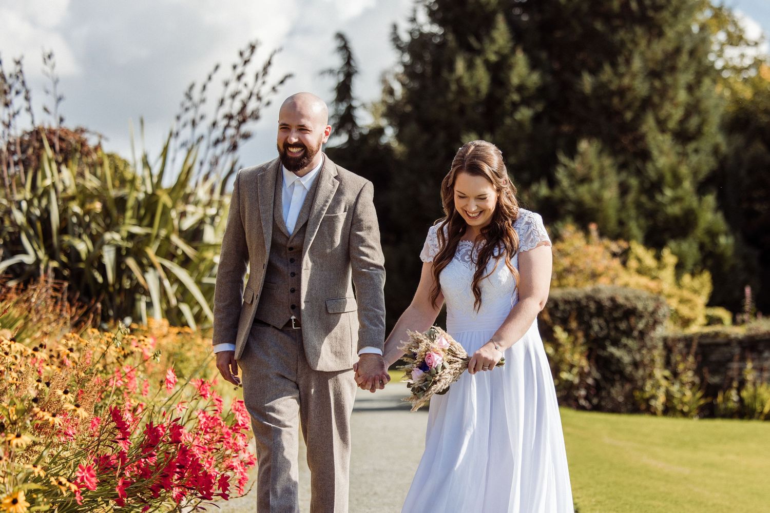A bride and groom walk hand in hand through a lush garden, she in a white gown, he in a tan suit.