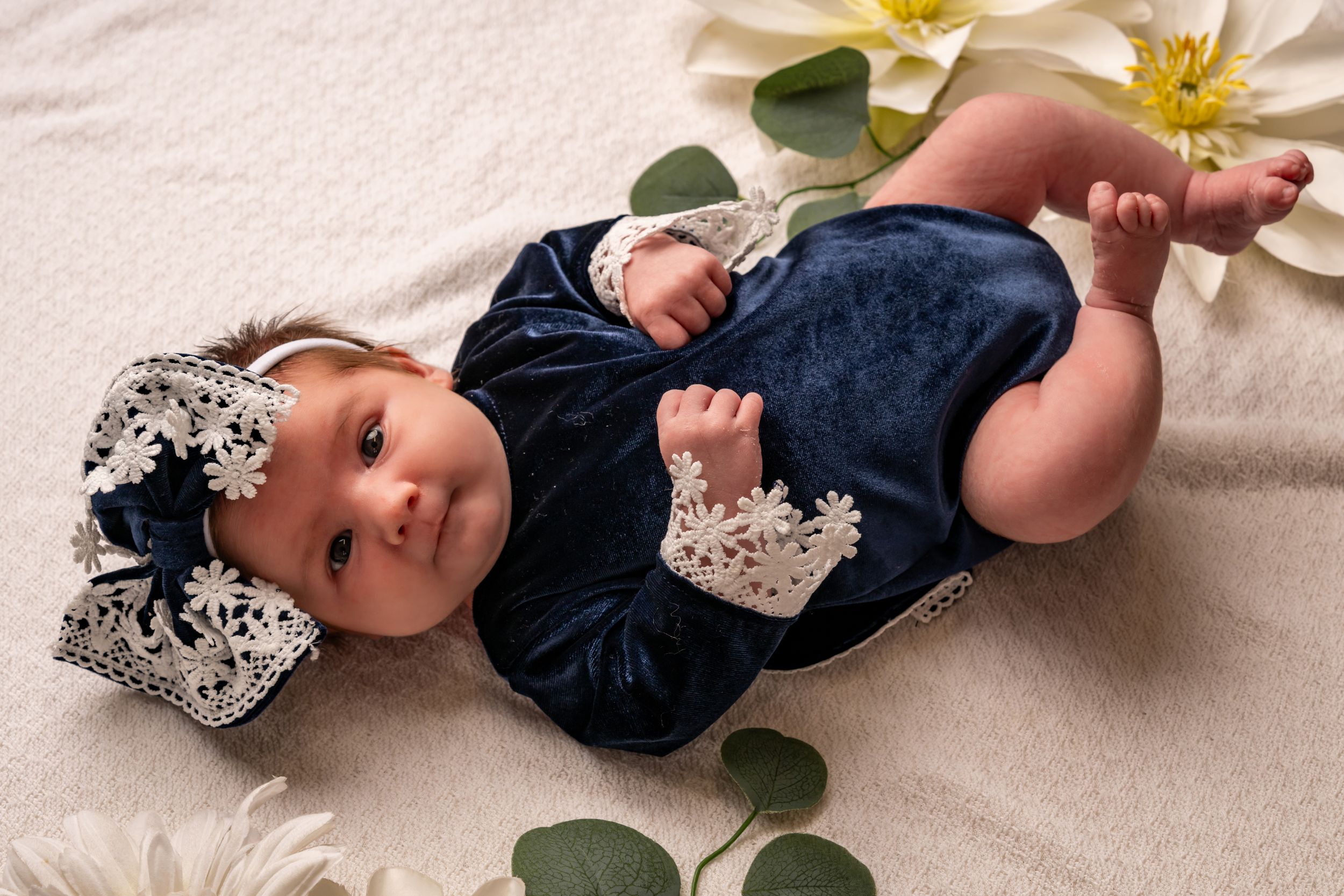 Awake newborn wearing a navy outfit and matching bow with white lace, lying on a cream blanket