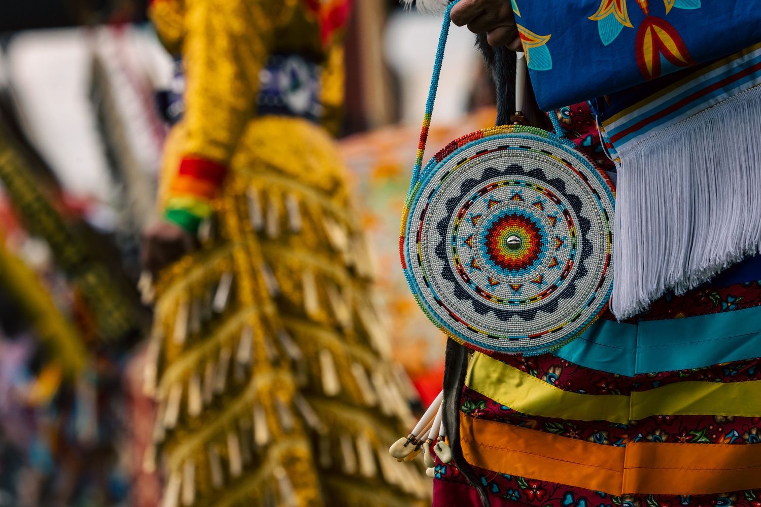 Close-up of a round beaded medallion bag with fringe, worn with colorful regalia and a feathered dancer in the blurred background.