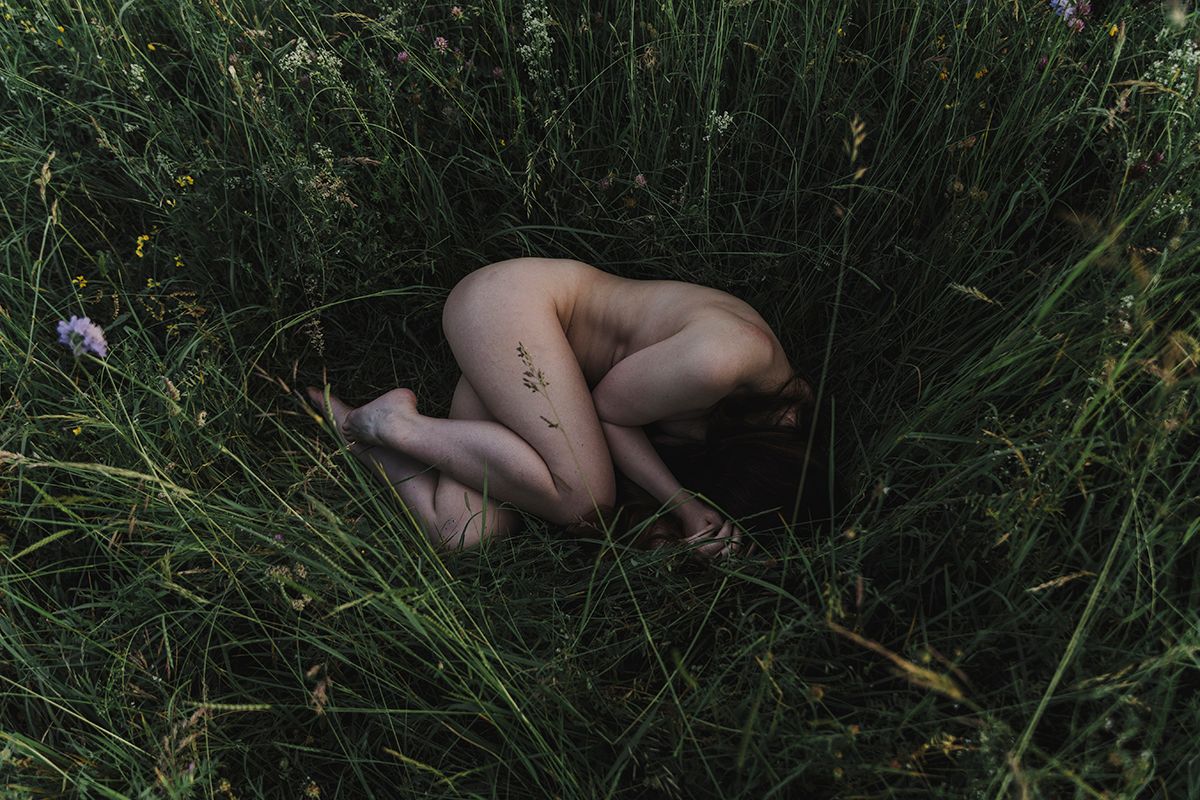 Person curled up in a fetal position among tall green grass and wildflowers, photographed from above.