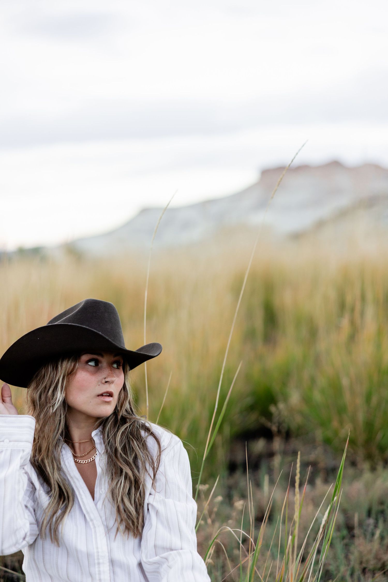 Woman in a black cowboy hat and white striped shirt looks to the side in a golden grass field with a rugged mesa in the background.