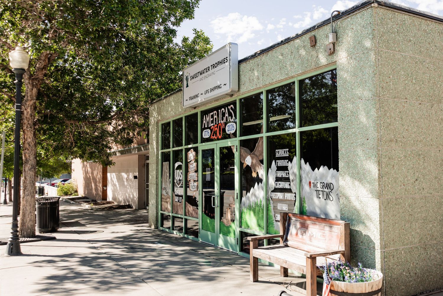 Green storefront with large windows and a hanging sign on a tree-lined sidewalk, with a wooden bench and flower planter out front.