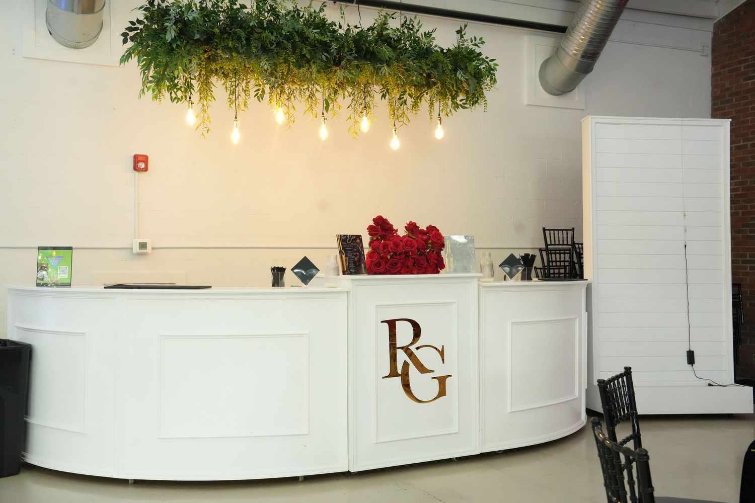 White reception desk with RG logo, adorned with red roses and hanging greenery with Edison bulbs above.