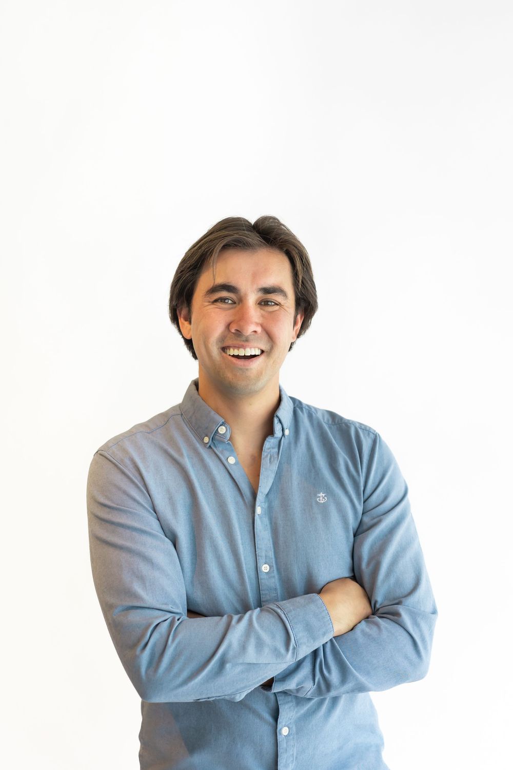 Smiling man with dark hair, arms crossed, wearing a blue button-up shirt, posing against a white background.