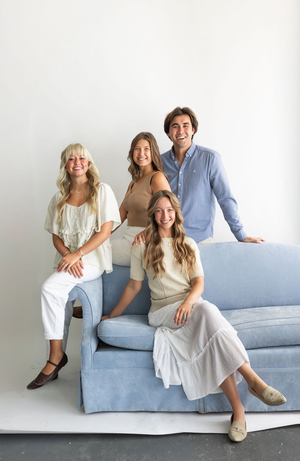 Four smiling young adults posing together on a light blue sofa in a bright studio setting.