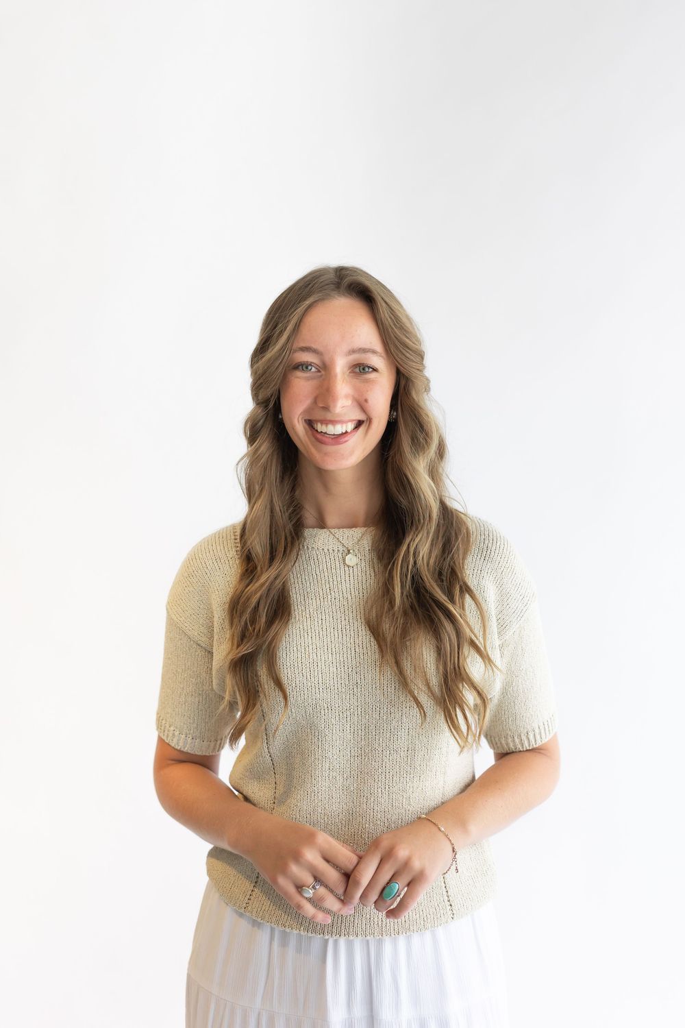 Smiling young woman with long wavy hair wearing a beige knit top and white skirt against a white background.