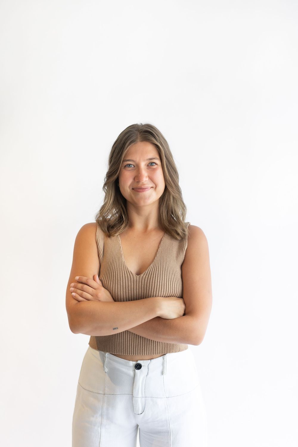 Young woman in beige ribbed tank top and white jeans, arms crossed, smiling against white background.