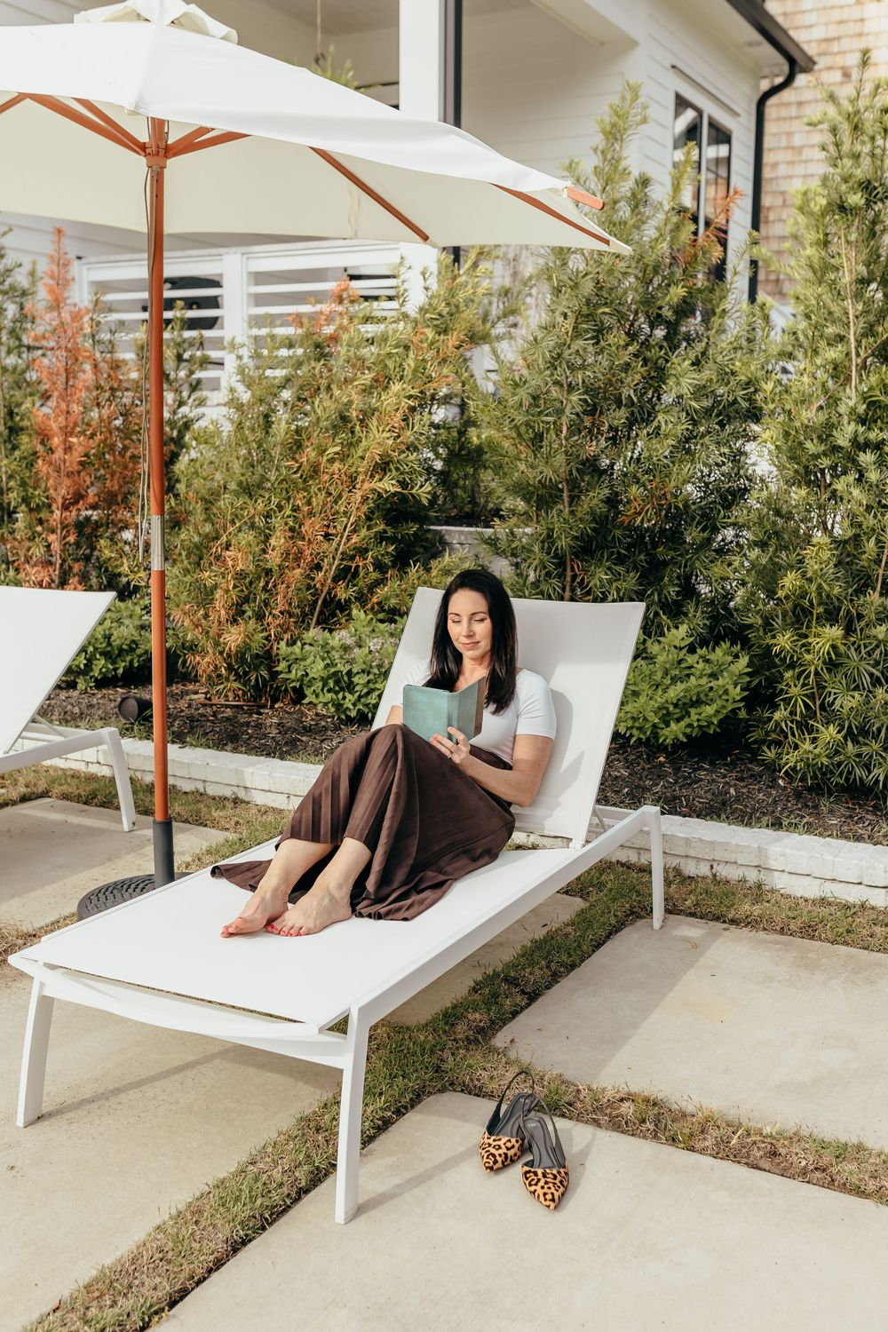 Woman relaxing on a white lounge chair outdoors, reading a book under a patio umbrella beside lush green hedges.