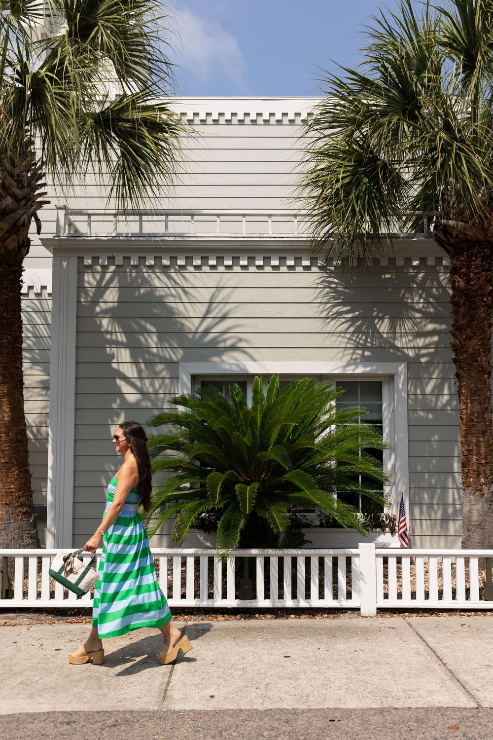 Woman in green striped maxi dress walking past a white building with palm trees on a sunny day.