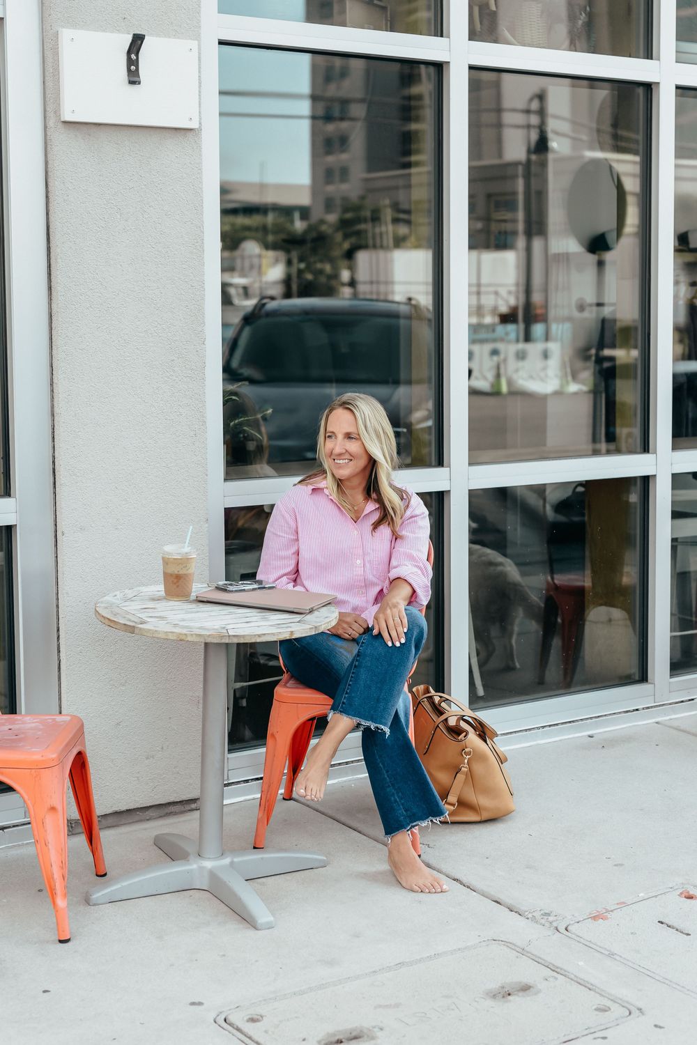 Woman in pink shirt and jeans sitting at outdoor cafe table with iced coffee and tan handbag.