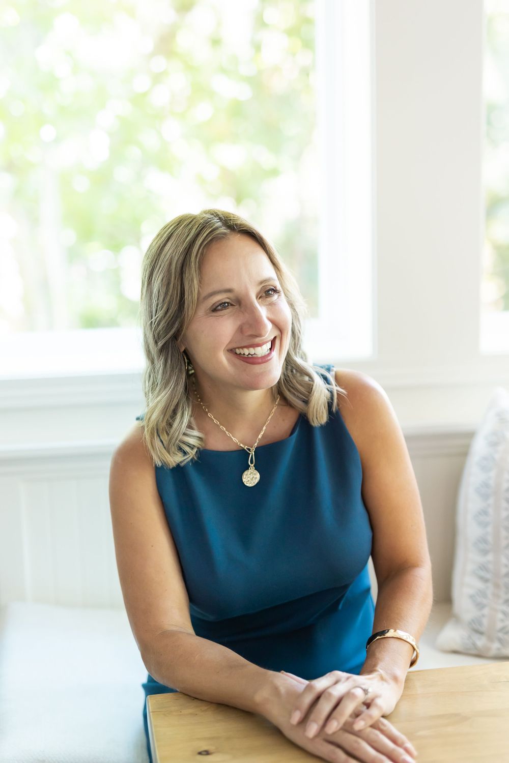 Smiling blonde woman in teal sleeveless dress wearing gold necklace, seated at wooden table near bright window.