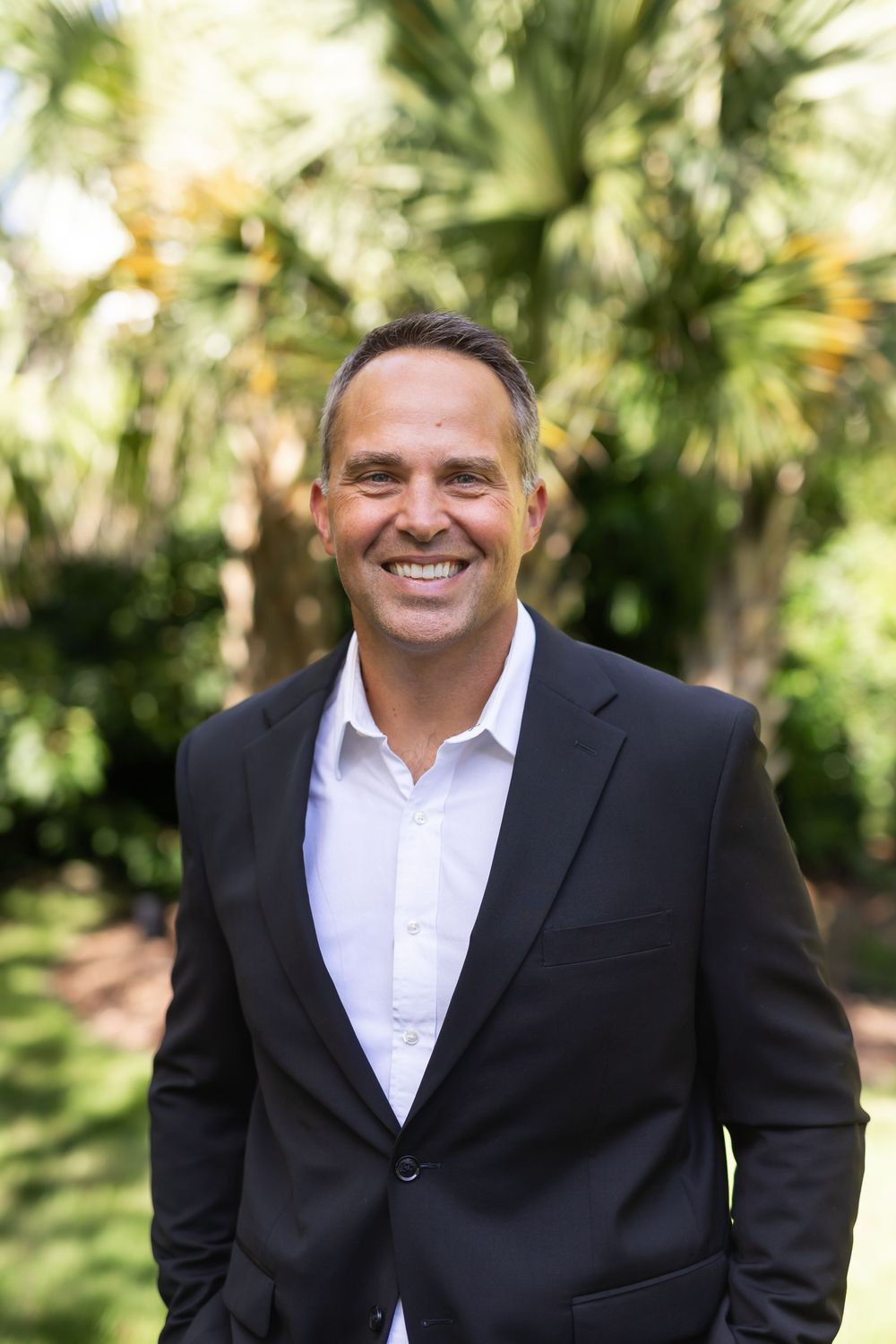 Smiling man in black blazer and white shirt standing outdoors among lush green palm trees.