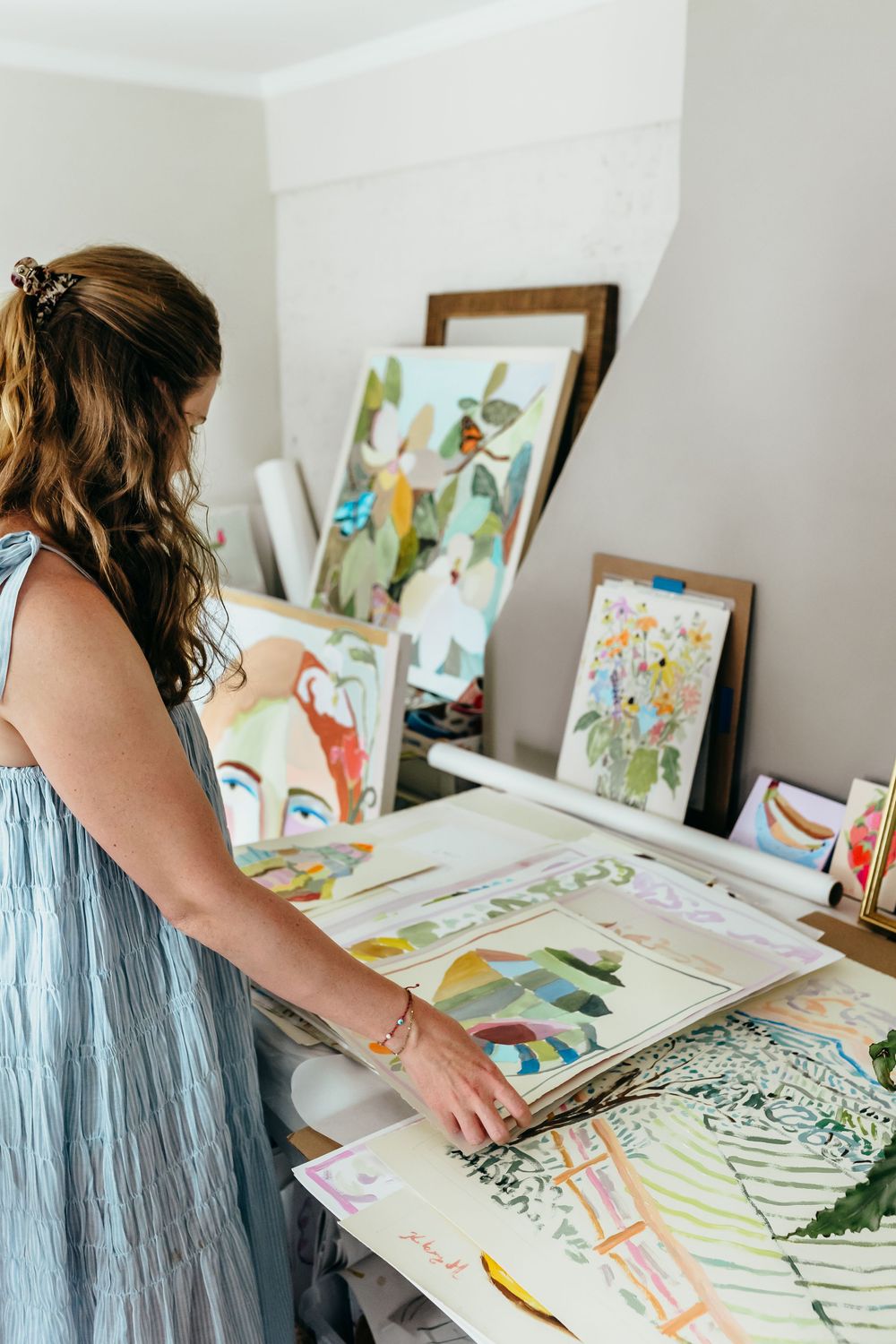 Artist in blue dress reviewing colorful paintings and artwork in a bright studio workspace.