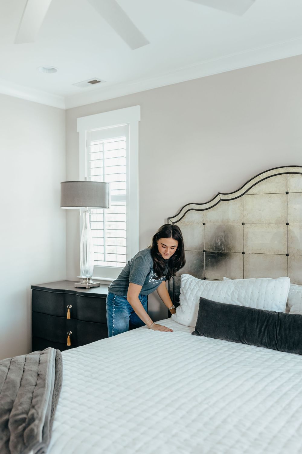 Woman making bed with white quilted bedding in elegant bedroom with tufted headboard.