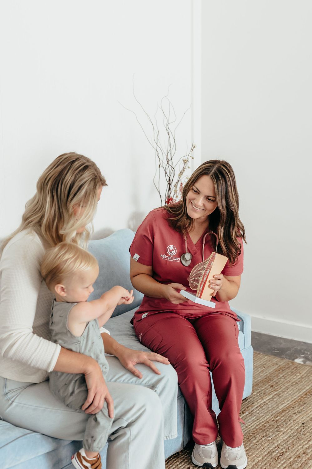 A nurse in red scrubs shows a brain model to a mother and toddler seated together in a bright medical office.