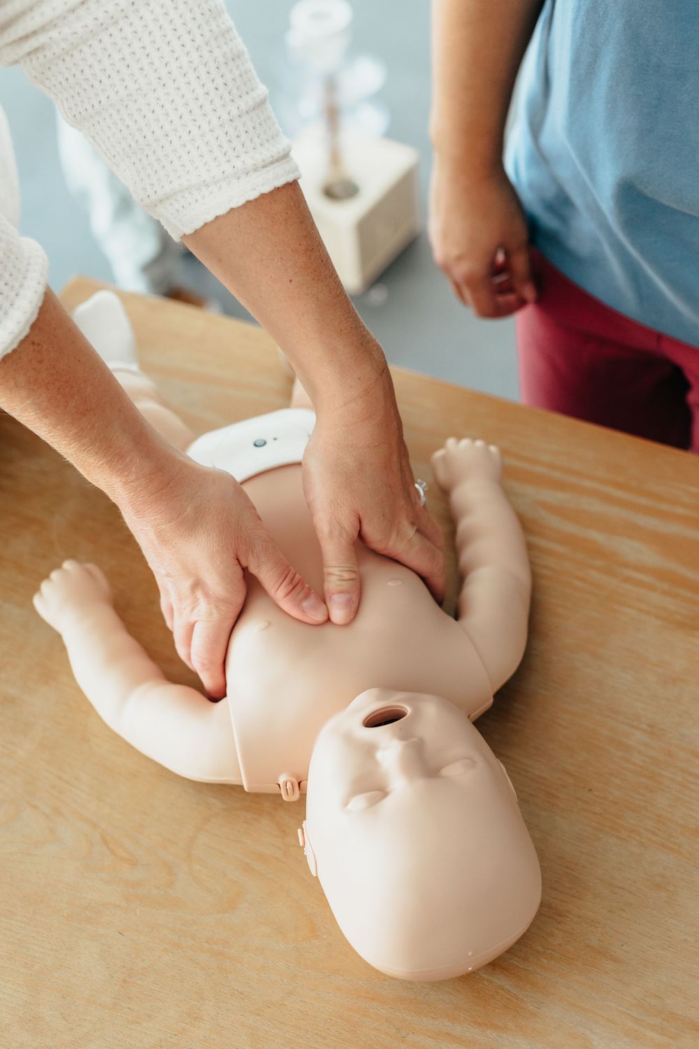 Hands performing CPR chest compressions on a infant CPR training manikin on a wooden table.
