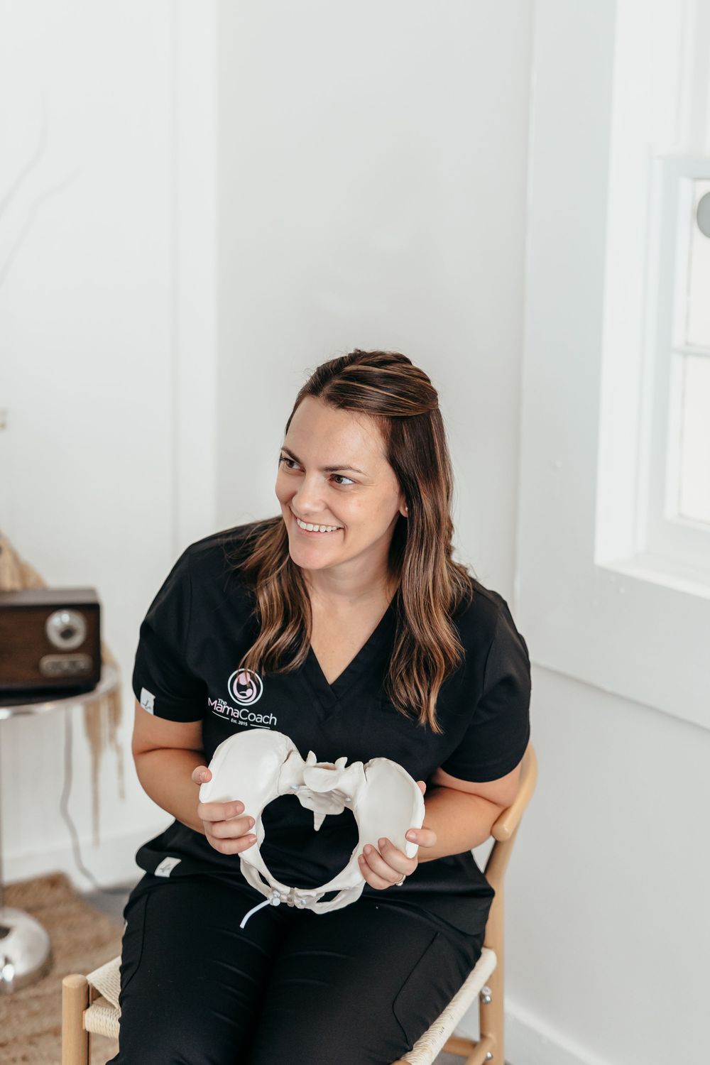 Smiling female healthcare professional in black scrubs holding a pelvis anatomical model while seated in a bright clinic.