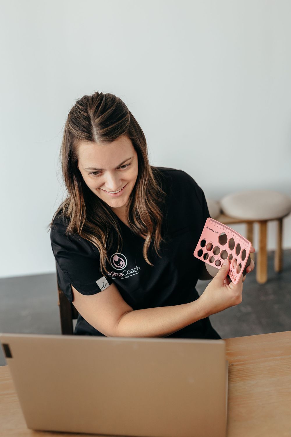 Woman in black branded shirt smiling at laptop while holding a pink product sample card at a desk.