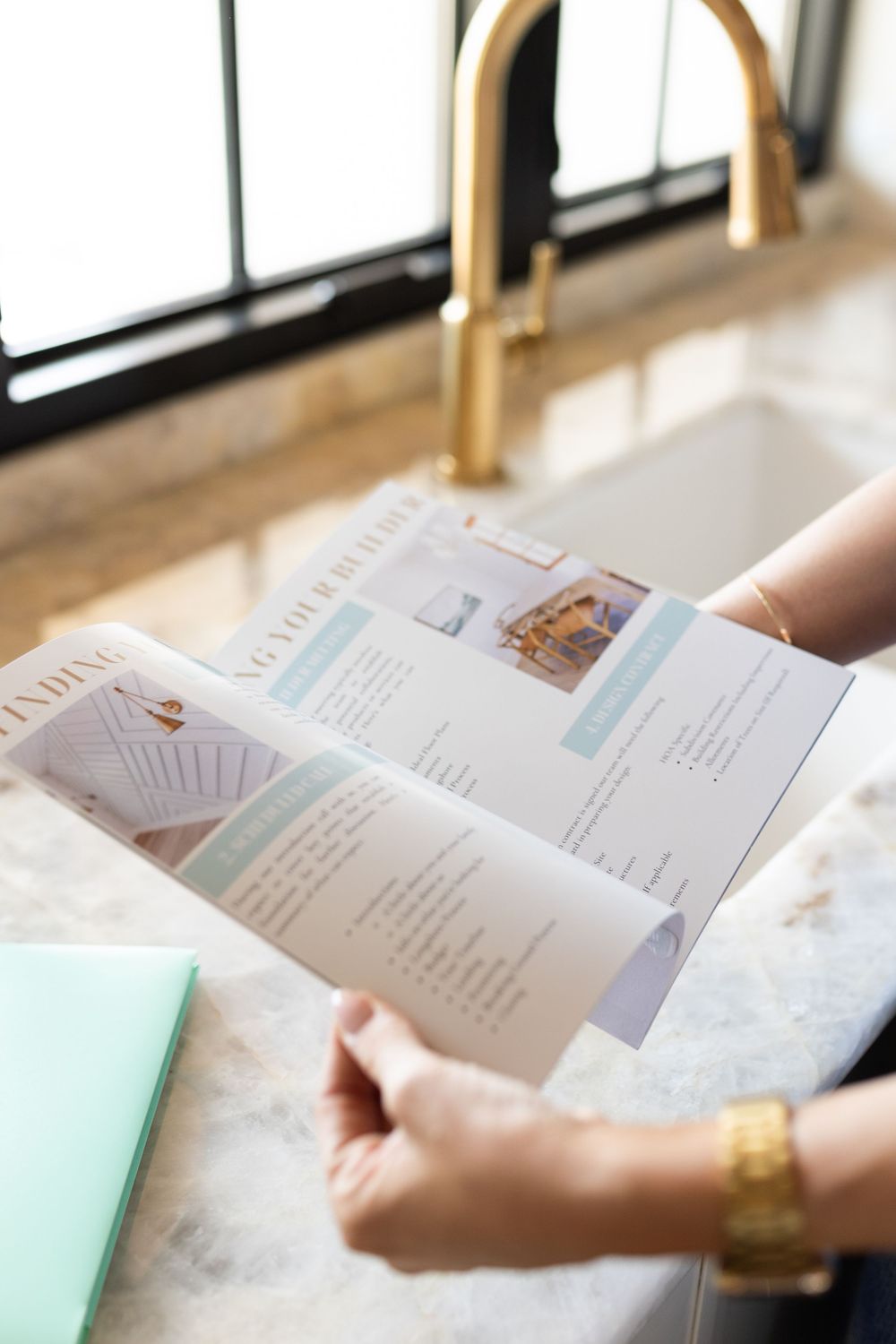 Person holding an open brochure near a marble countertop with a gold faucet in a modern kitchen.