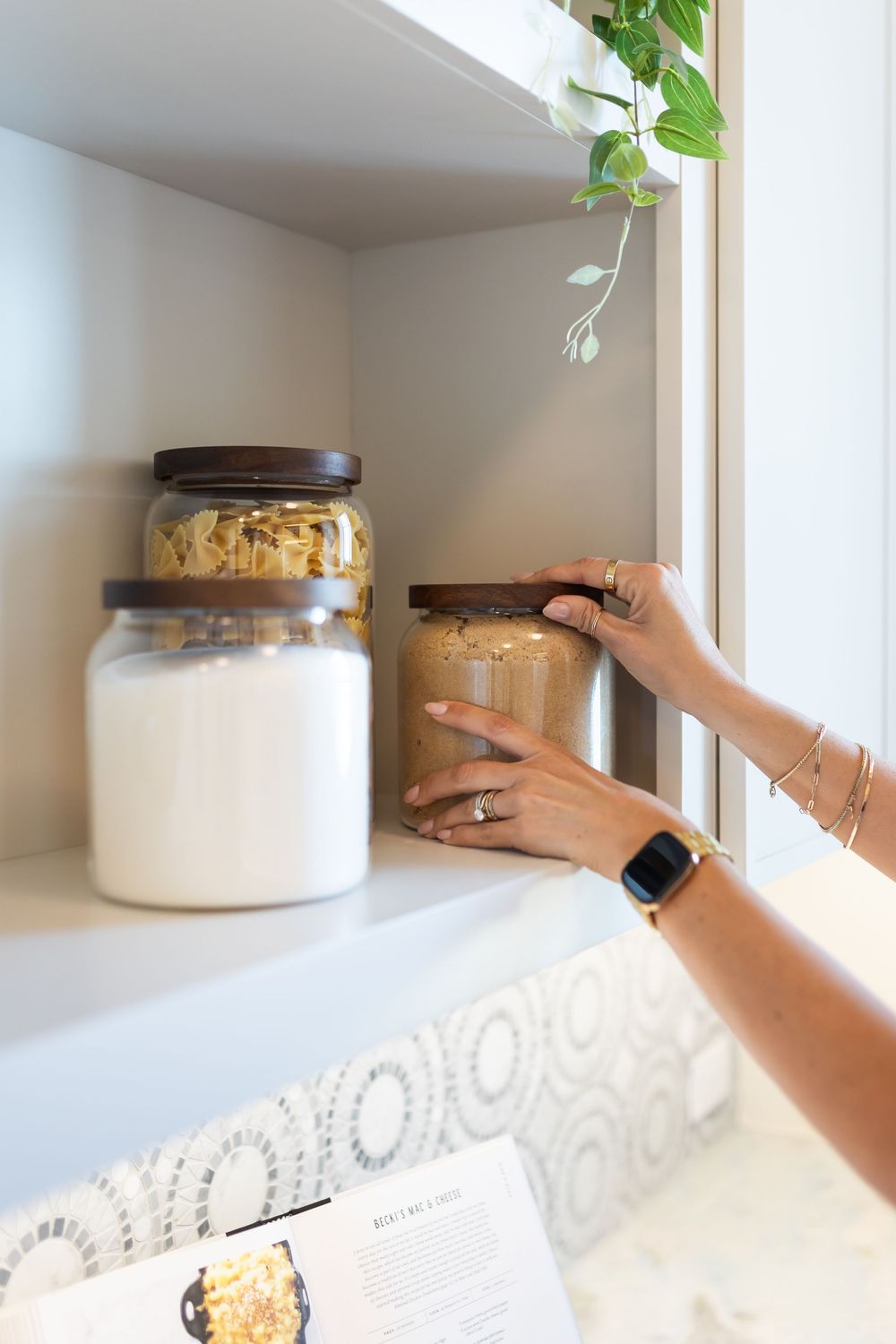 Woman placing a glass jar with brown lid on a white kitchen shelf beside flour and granola storage jars.