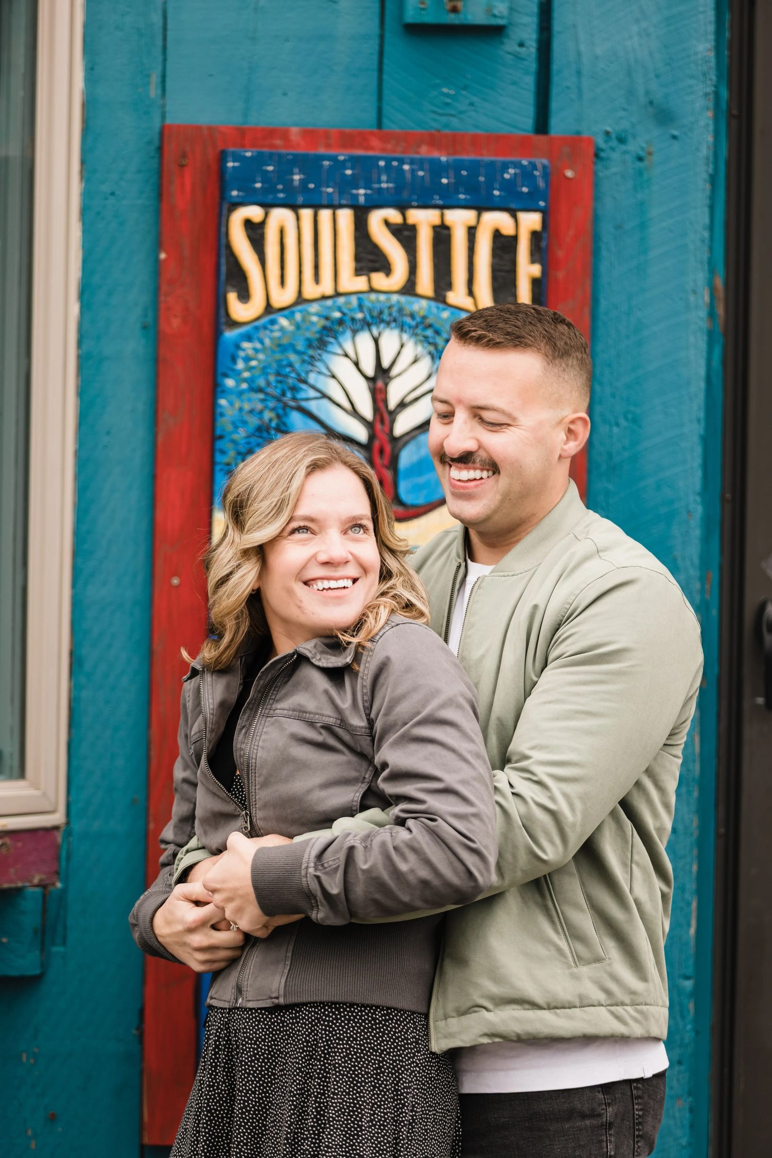 A couple embraces and smiles in front of a colorful mural featuring the word 'Soulstice' and a tree design. Engagement at Rockport Harbor,