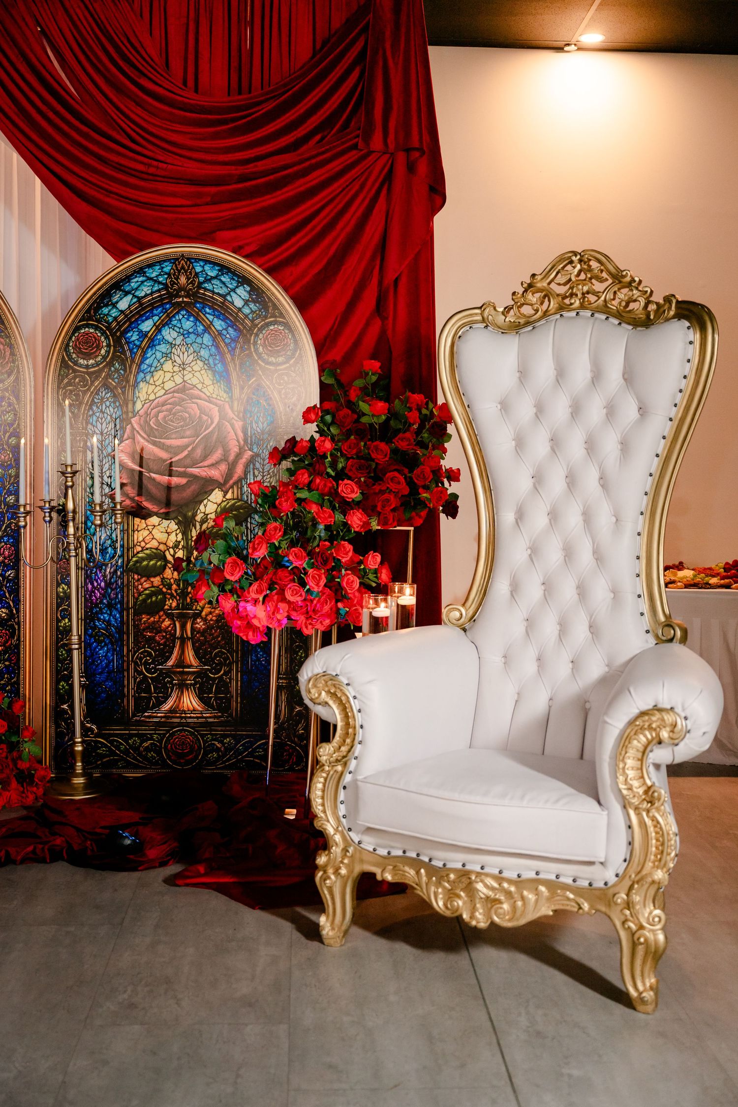 White tufted throne chair with gold trim beside red roses, a stained glass rose panel, and red velvet drapes at an elegant event.