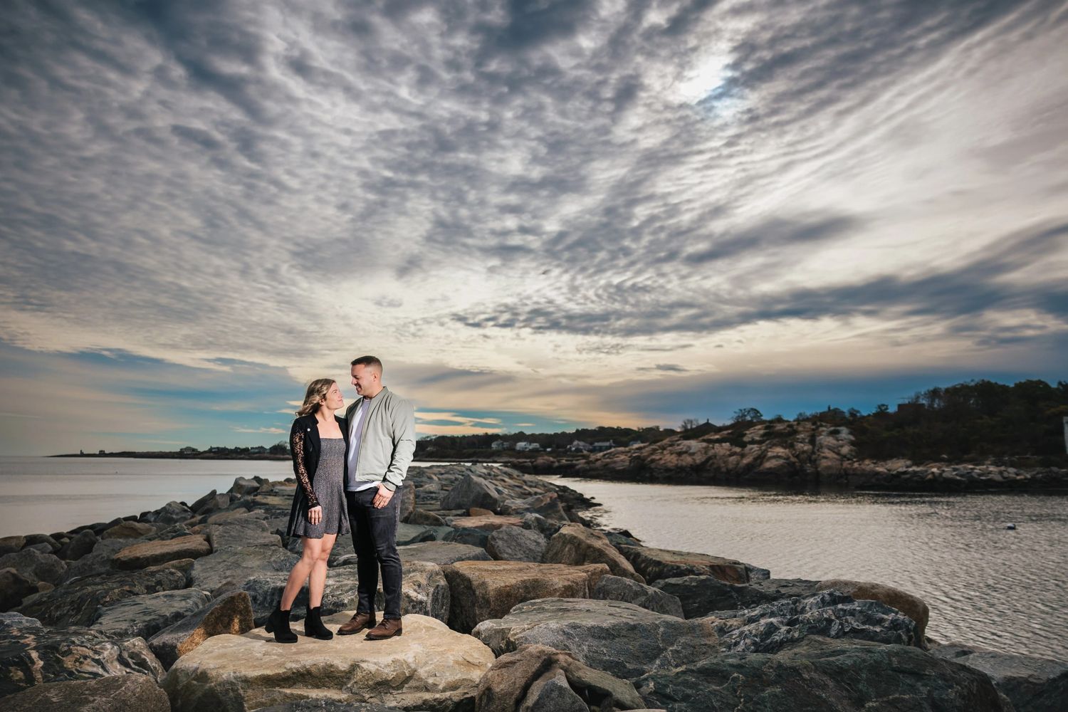 Couple standing together on stone breaker at dawn gazing toward the water. Engagement at Rockport Harbor, Rockport, MA