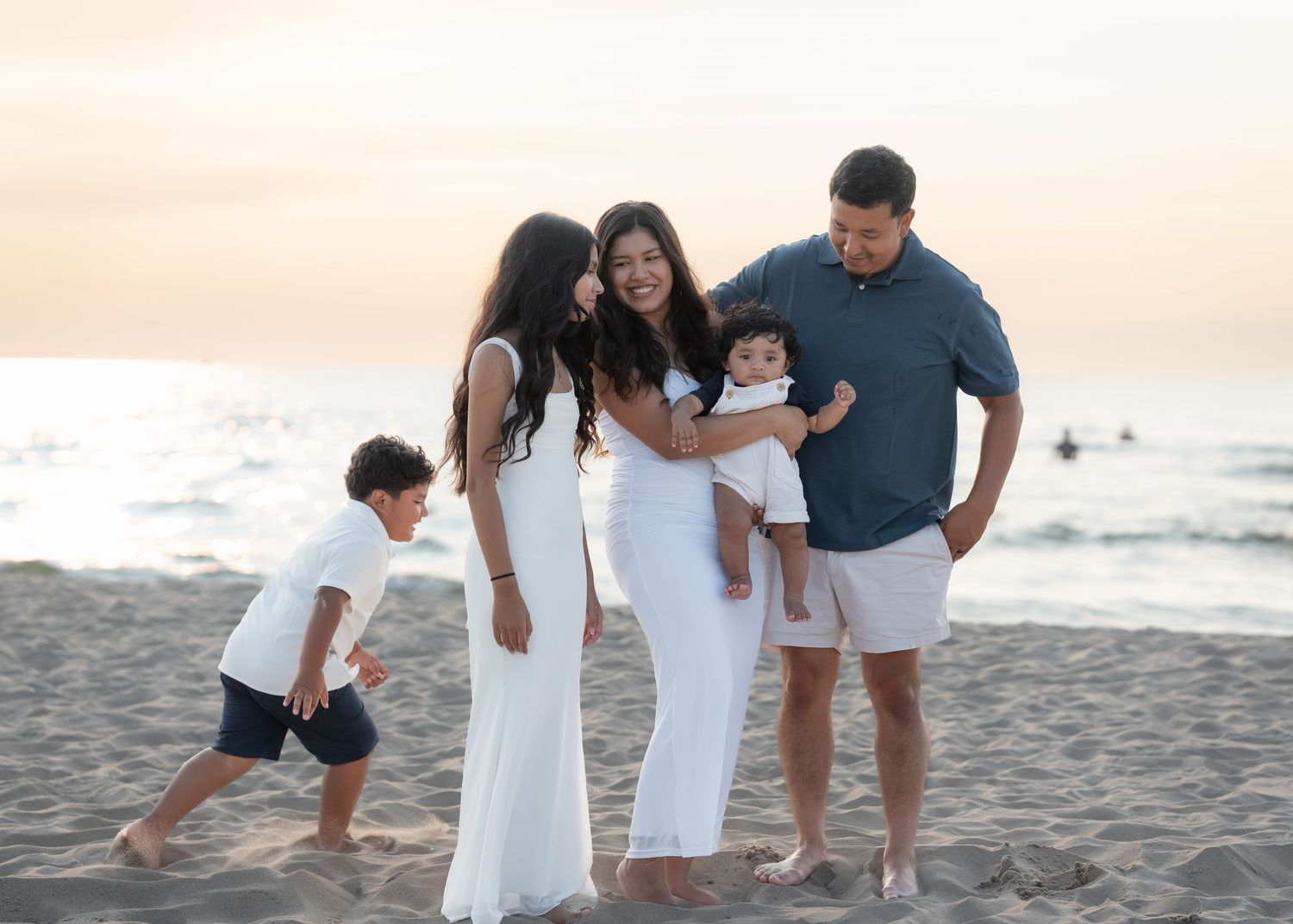 Happy family of five poses on a sandy beach at sunset, mother in white dress holding toddler.