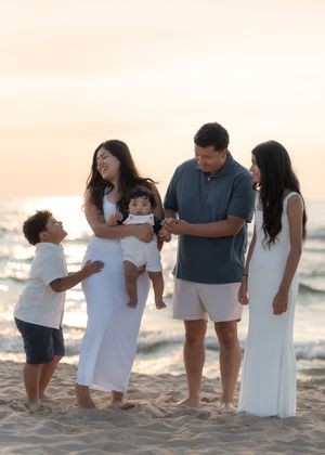 Family of five smiling and laughing together on a sandy beach at sunset, dressed in white and blue outfits.