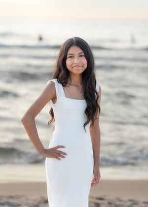 Young girl in white dress poses on beach at sunset with ocean waves in background.