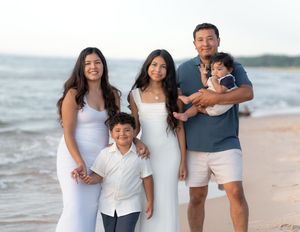 Family of five posing on a sandy beach, wearing white and blue outfits, with a man holding a baby.