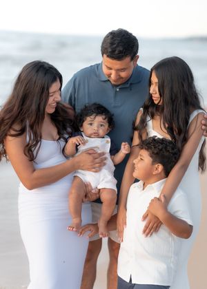 Family of five posing together on a beach, parents holding a baby while two children smile nearby.