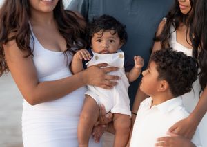 Mother in white dress holding infant while young boy stands beside her during family photo session.