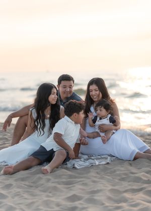 Family of five sitting on sandy beach at sunset, parents and three children in white outfits smiling together.
