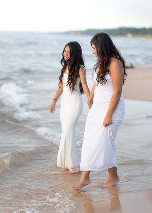 Two young women in white maxi dresses walk barefoot along a sandy beach shore with gentle waves.