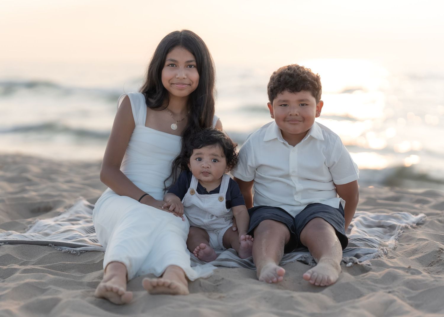 A woman and two young children smile together on a sandy beach at sunset in coordinating white outfits.