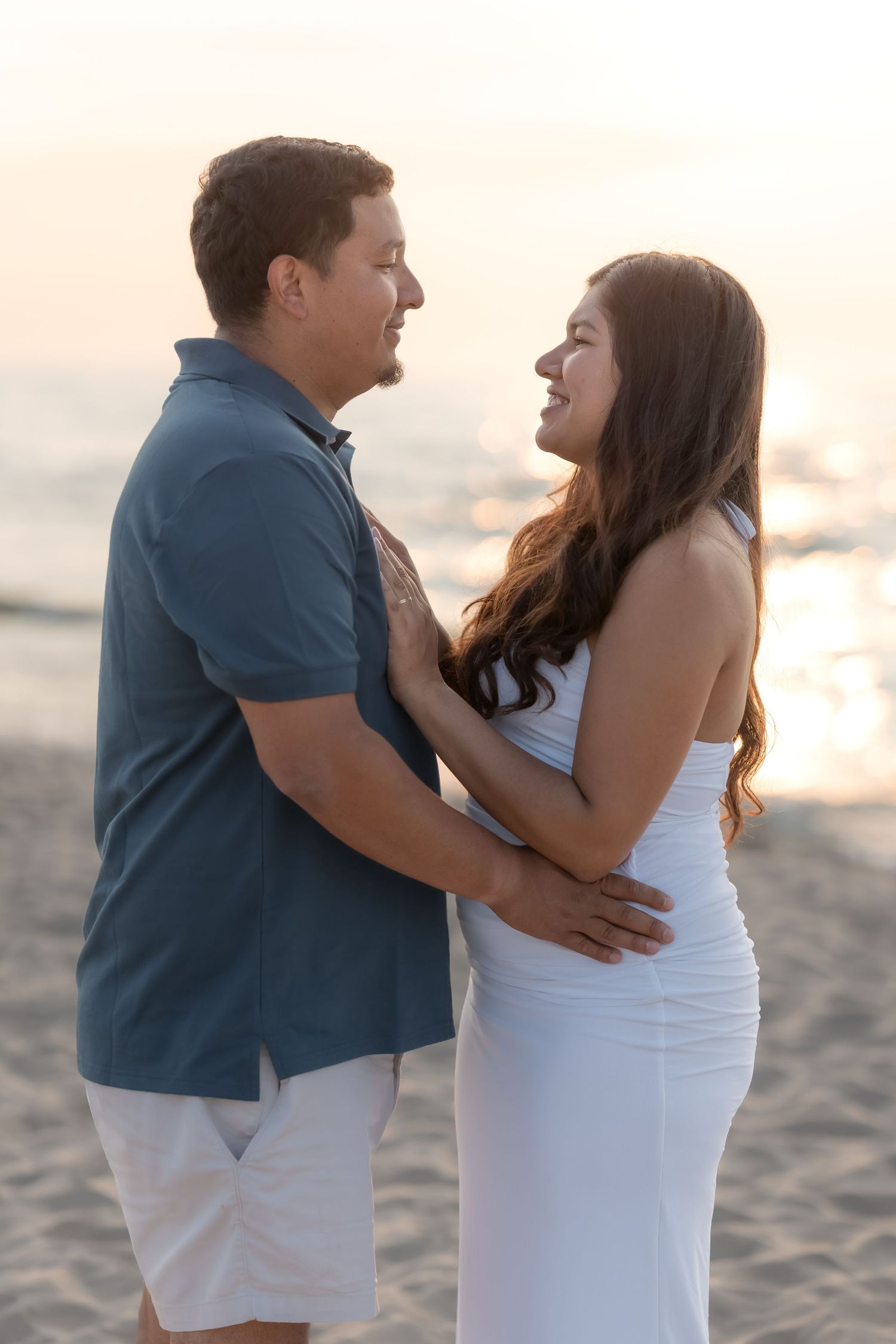 Couple embracing and smiling at each other on a beach at sunset, man in blue shirt, woman in white dress.