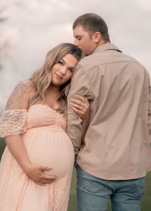 Pregnant woman in pink lace gown embraced by partner outdoors during romantic maternity photo session.