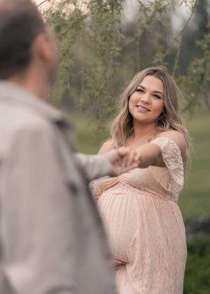 Pregnant woman in blush lace maternity gown holds hands with partner outdoors near weeping willow trees.